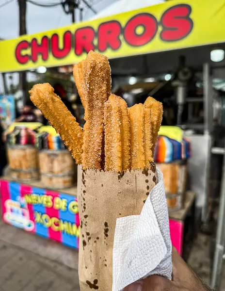 Golden-brown churros coated in sugar, standing upright in a paper cone with a napkin, held in front of a bright yellow street-food stand sign that reads “CHURROS,” with colorful market stalls blurred in the background.