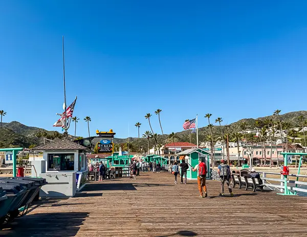 Visitors walking along the Green Pleasure Pier in Avalon, Catalina Island, with shops, ticket booths, and American flags visible.