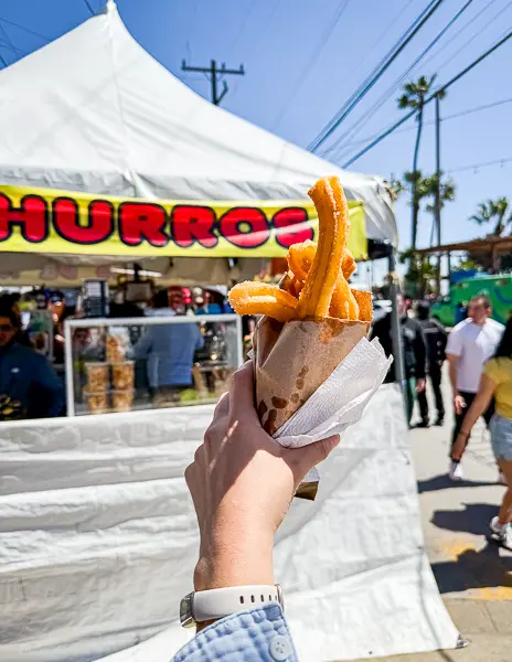 Hand holding a paper cone of cinnamon sugar churros in front of a street market stand with a yellow “Churros” sign in Ensenada, Mexico.