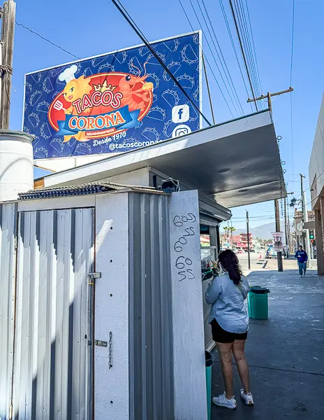 Exterior of Tacos Corona taco stand in Ensenada, Mexico, with the restaurant sign above and Kathy ordering at the counter.