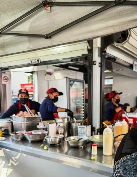 Workers preparing fish and shrimp tacos at the Tacos Fenix stand in Ensenada, Mexico, with fresh toppings and sauces on the counter.