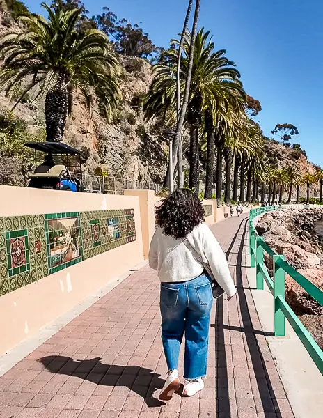 Person (kathy) walking along Casino Way in Avalon, Catalina Island, with palm trees, ocean views, and colorful tile walls lining the path toward the Casino.
