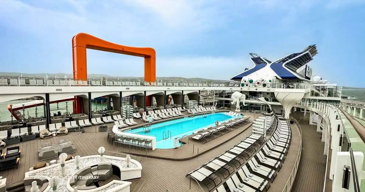featured image: The main pool deck aboard a Celebrity Cruises Edge Series ship featuring the signature orange Magic Carpet platform, lounge chairs, and modern design under a clear blue sky.