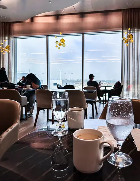 Coffee mugs and water glasses on a dark marble table inside an airport lounge, with floor-to-ceiling windows and travelers seated in soft chairs overlooking the runway.