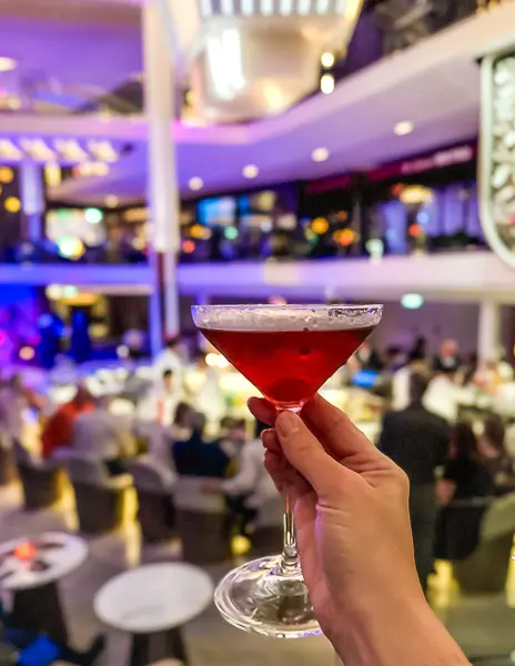 Hand holding a martini at the Martini Bar on a Celebrity Cruise ship, with the lively bar and lounge in the background.