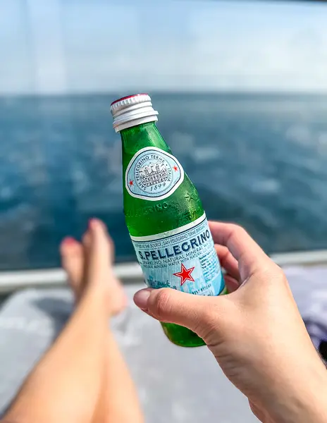 Hand holding a chilled San Pellegrino sparkling water on a Celebrity Cruise balcony with the ocean in the background.