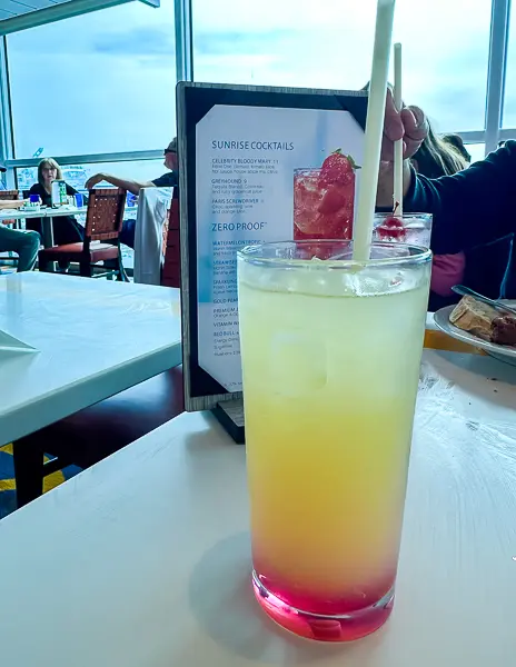 Colorful zero proof cocktail served on a Celebrity Cruise ship, with the bar menu showing Sunrise Cocktails and Zero Proof options in the background.