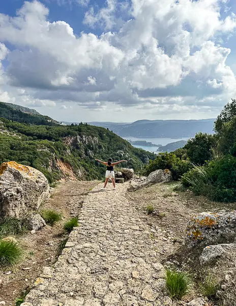 Standing on a rocky path overlooking the lush green hills and coastline near Angelokastro, a historic hilltop castle in Corfu, Greece — reached during a private driver tour on a Mediterranean cruise port day.