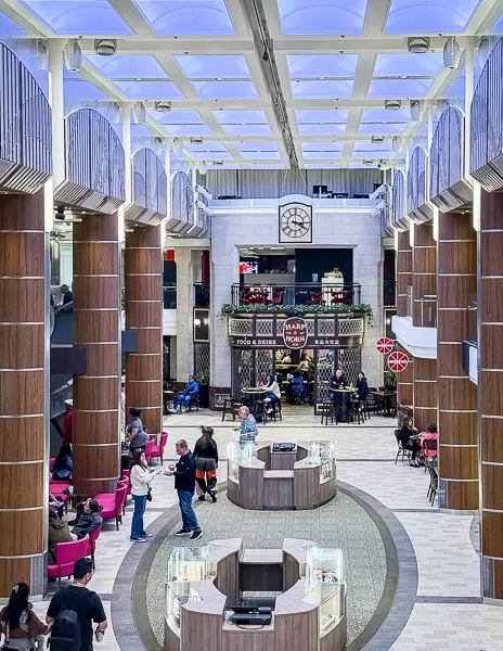 The Royal Esplanade on Royal Caribbean’s Quantum of the Seas, with passengers walking through the bright indoor walkway lined with shops, seating areas, and La Patisserie at the far end. Display cases are set up in the center for ongoing sales on Deck 4.