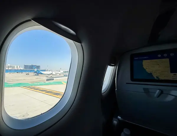 Looking out the airplane window while seated near the front of the cabin, with the in-flight map on the seatback screen and Alaska Airlines planes visible on the tarmac at LAX.