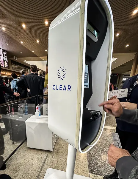 CLEAR verification kiosk at LAX airport with travelers lined up behind and a staff member assisting with the process.