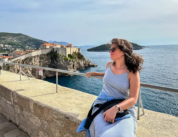 Woman (Kathy) sitting along Dubrovnik’s historic city walls with the Adriatic Sea and Fort Lovrijenac in the background during a Game of Thrones-themed walking tour in Croatia.
