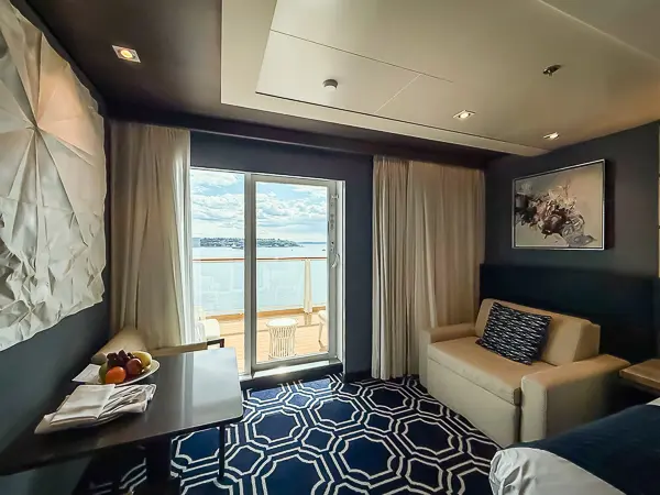 Interior of a Haven Penthouse suite on the Norwegian Encore, showing a modern sitting area and balcony with an ocean view through the sliding glass doors. A bowl of fruit sits on the nearby table.