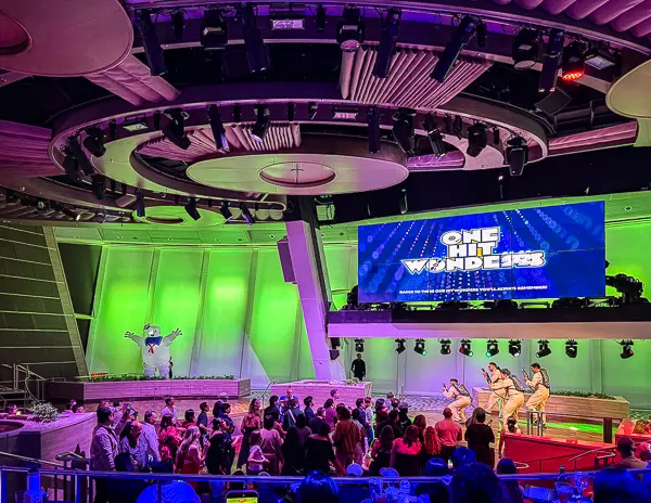 Guests gathered in Two70 on Royal Caribbean’s Quantum of the Seas during the “One Hit Wonders” party, with performers on stage, colorful lighting, and a large screen displaying the event name.
