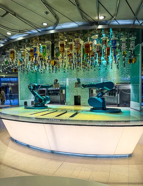 Full view of the Bionic Bar on Quantum of the Seas, showing both robot bartenders and the overhead liquor system illuminated in teal lighting.