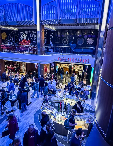 View of Café Promenade on Royal Caribbean’s Quantum of the Seas, with passengers browsing merchandise tables and enjoying the busy indoor Esplanade area under blue lighting