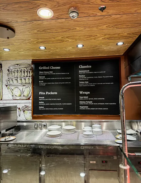 Menu board inside Café Two70 on Quantum of the Seas, listing grilled cheese options, pita pockets, classics, and wraps above the service counter.