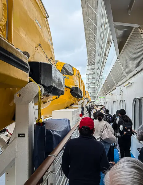 Passengers lined up along the outdoor deck of Quantum of the Seas during debarkation, walking past the ship’s yellow lifeboats toward the exit on a gray morning.