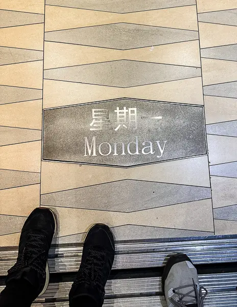 “Monday” displayed on the elevator floor of Quantum of the Seas, with the day spelled out in English and Chinese characters, and two passengers’ shoes visible at the edge of the frame.