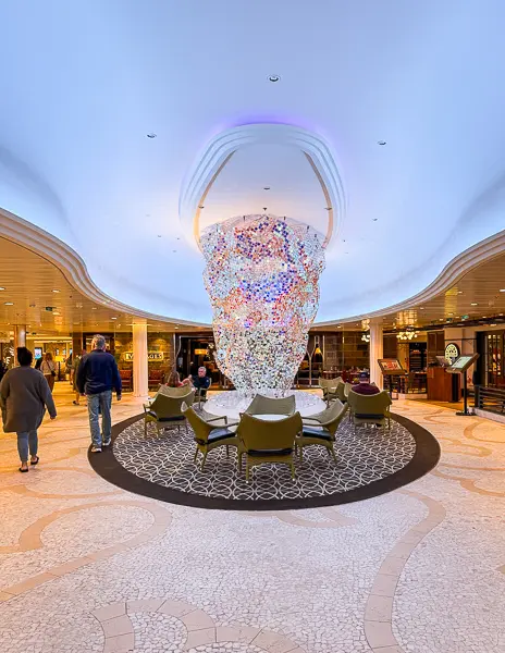 A view of the Royal Esplanade on Quantum of the Seas, featuring the large multi-colored crystal installation at the center of a circular seating area. Guests walk through the bright, open space with shops and venues visible in the background.