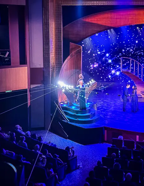 Scene from Sonic Odyssey on Quantum of the Seas, with a performer playing the Earth Harp on stage as beams stretch across the theater and the audience watches.
