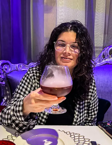 Kathy holding the Smoked Rings cocktail at Wonderland on Quantum of the Seas, seated at a table with the restaurant’s purple lighting in the background.