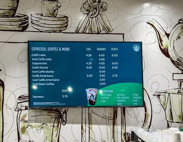 Digital menu board displaying Starbucks-style espresso and coffee drinks inside Café Two70 on Royal Caribbean’s Quantum of the Seas. Prices for lattes, cappuccinos, mochas, and americanos are shown in Tall, Grande, and Venti sizes, with add-on options listed at the bottom.