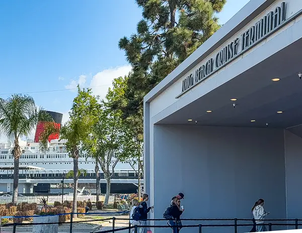 Entrance to the Long Beach Cruise Terminal with passengers walking in and the Queen Mary visible behind the trees.