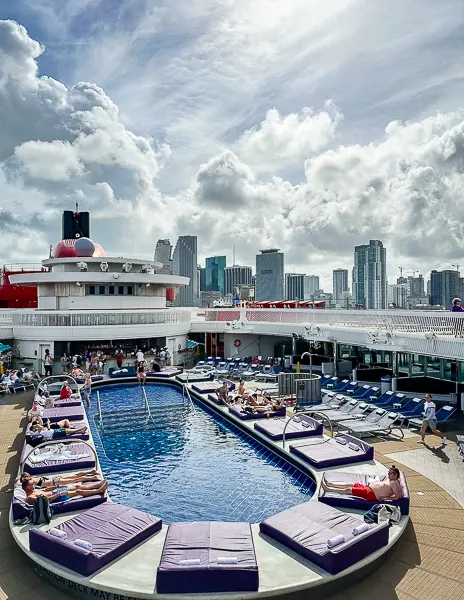 View of the Virgin Voyages pool deck with loungers and the Miami skyline in the background, highlighting that certain cruise lines and newer ships may only sail from specific homeports.