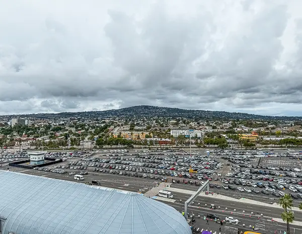 View of the parking lot at the San Pedro World Cruise Center, with rows of cars, the terminal roof, and hillside neighborhoods in the background on a cloudy day.