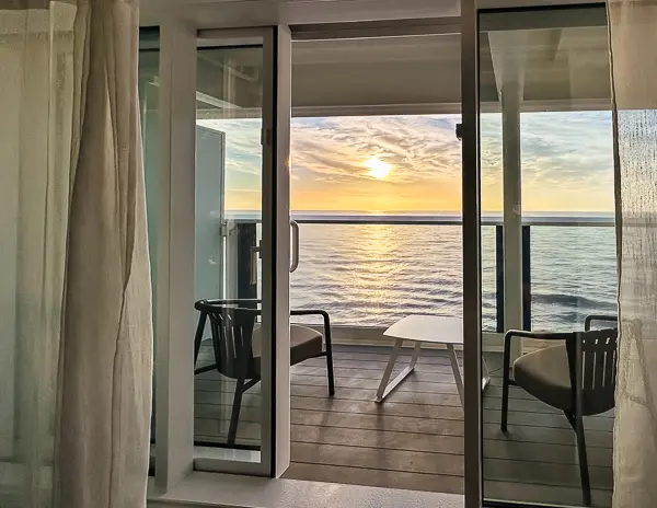 Sunrise over the Mediterranean Sea viewed from a cruise ship balcony, with open balcony doors, chairs, and calm water stretching to the horizon.