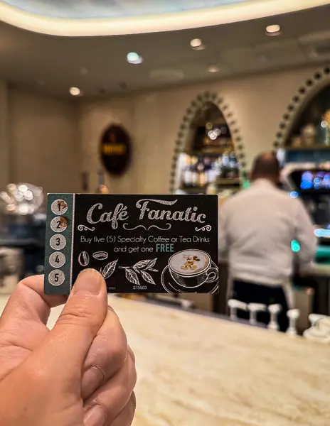 Hand holding a Disney cruise coffee rewards card at an onboard café, showing buy five specialty coffee or tea drinks and get one free.