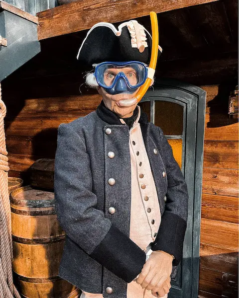 A historical maritime figure in period clothing wears a modern snorkel mask while standing inside a wooden ship cabin, part of Norwegian Cruise Line’s new social media campaign.