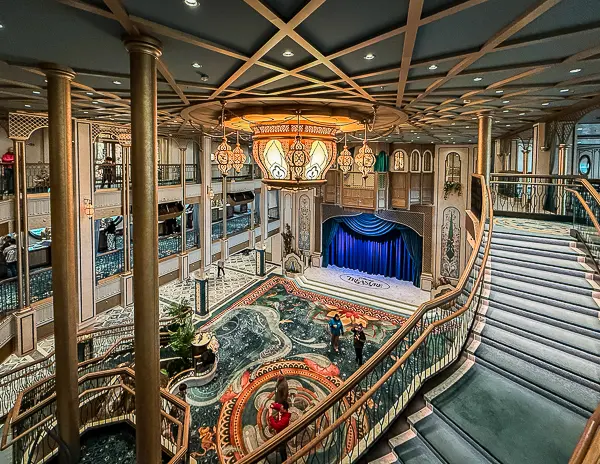 Grand Hall on Disney Treasure featuring sweeping staircases, ornate chandelier, patterned carpet, and stage with blue curtains.