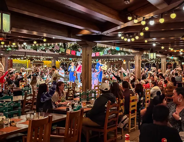 Lively dinner show inside Plaza de Coco on Disney Treasure with performers on stage and guests dining at long wooden tables.