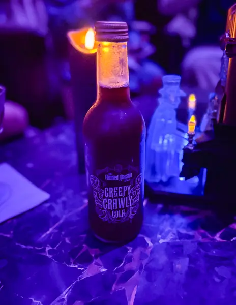 Creepy Crawly Cola bottle on the bar inside the Haunted Mansion Parlor on Disney Treasure, surrounded by dim purple lighting and spooky themed décor.