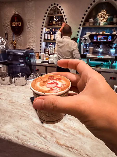Hand holding a macchiato with Hei Hei latte art at Hei Hei Café on Disney Treasure, with the barista and espresso machines in the background.