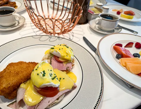 Eggs Benedict served at 1923 on Disney Treasure with hollandaise sauce, sliced ham, and a hash brown on the side, alongside fruit and coffee.