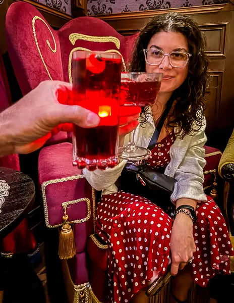 Hubby and I clinking red cocktails inside the Haunted Mansion Parlor on Disney Treasure, seated in themed red velvet chairs with gothic décor.