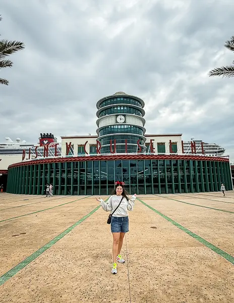 Woman (Kathy) wearing Minnie ears standing in front of the Disney Cruise Line terminal at Port Canaveral with the Disney ship visible behind her under cloudy skies.
