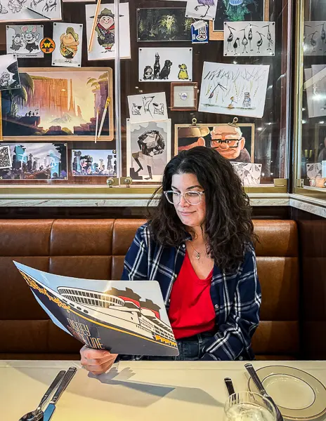 Kathy seated at 1923 restaurant on Disney Treasure reviewing the dinner menu, with Disney animation sketches displayed on the wall behind her.