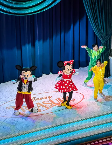 Mickey Mouse and Minnie Mouse performing on stage during a Disney Treasure show, dancing alongside two performers in colorful costumes.