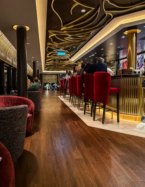 Interior of The Rose lounge on Disney Treasure with red velvet bar stools and elegant gold ceiling details.