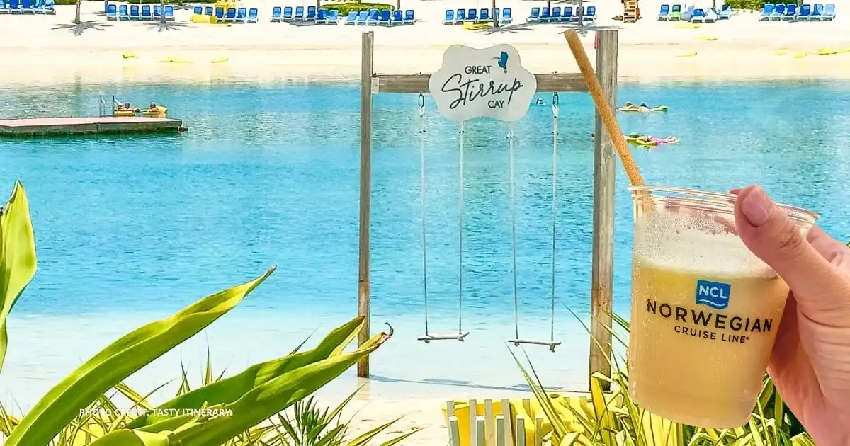 Great Stirrup Cay beach in the Bahamas with turquoise water, white sand, and a swing set in the background, while a hand holds a Norwegian Cruise Line drink cup in the foreground.