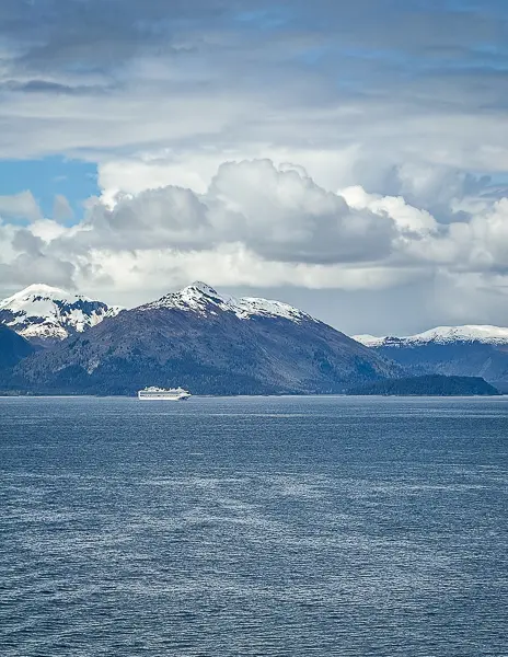 Cruise ship sailing past snow capped mountains and deep blue water along the Alaska coast under dramatic clouds.