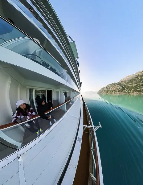 Kathy and her husband standing on their cruise ship balcony while sailing past calm water and mountains in Alaska.