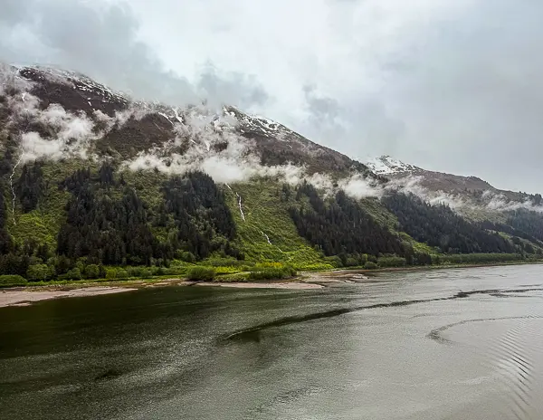 View from an Alaska cruise ship before docking in Juneau, with calm water, green shoreline, misty mountains, and low clouds.