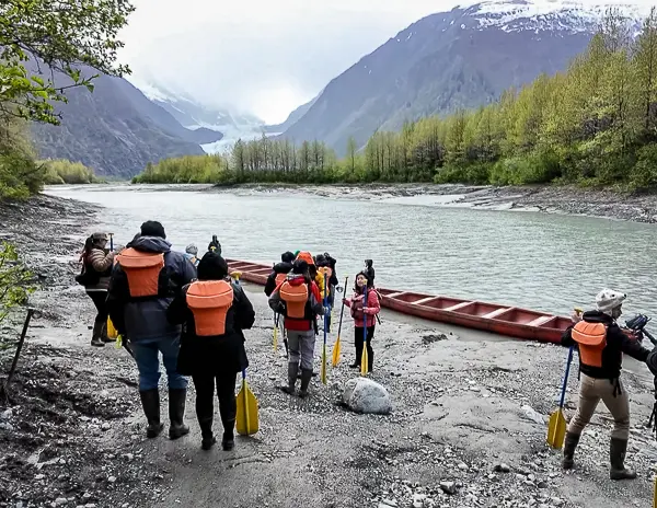 Tour guests / cruiser passengers standing near large canoes by glacier-fed water with Davidson Glacier visible in the distance.