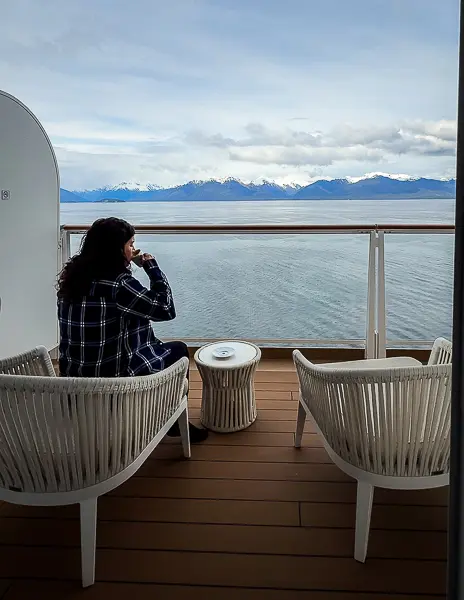 Woman (kathy) drinking coffee on an Alaska cruise balcony while looking out at calm water and snow-covered mountains.