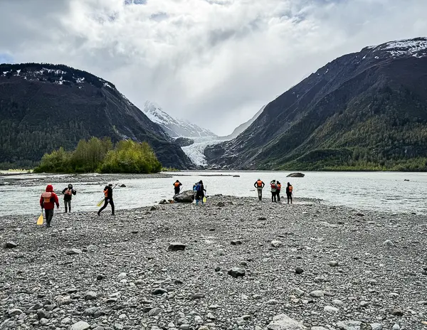 Tour guests / cruise passengers walking along the rocky shoreline with Davidson Glacier in the distance during a Skagway glacier excursion.